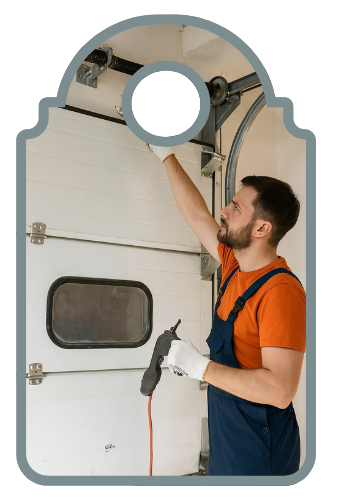 A garage door technician in orange and navy working on a white sectional door spring using a wrench and holding a drill inside a residential garage.