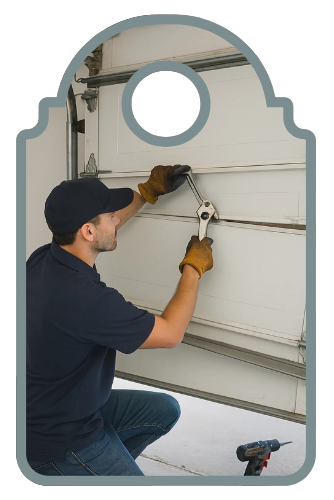  A garage door technician in a navy uniform installing a wood-finish sectional garage door on a residential property using a power drill, with beige siding and brick in the background.